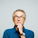 Portrait of elderly man wearing white denim shirt and glasses looking up with hand on chin. Thoughtful senior entrepreneur, studio shot against grey background.