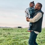 Black father holding daughter in a field of cows