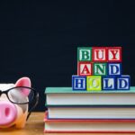 BUY AND HOLD spelled in letters on top of a pile of books. Alongside is a piggy bank in glasses. Buy and hold is a popular long term stock and shares strategy.